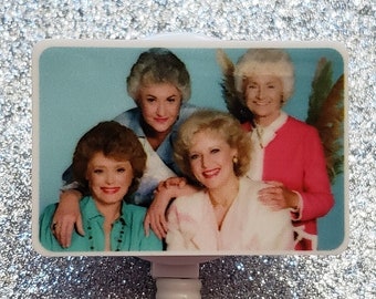 May include: A white retractable badge holder with a rectangular image of four women. The image shows the women smiling and posing together. The image is on a blue background.