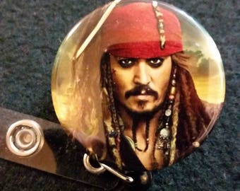 May include: A round button featuring a close-up image of Jack Sparrow from the Pirates of the Caribbean movie franchise. He is wearing a red bandana and has long brown hair and a beard. The background is a blurry image of a beach.