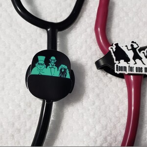 May include: Two black stethoscope charms. One charm features a green silhouette of three ghosts from the Haunted Mansion ride. The other charm features a black silhouette of three ghosts with the text "Room for one more?" in white.