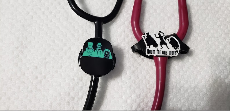 May include: Two black stethoscope charms. One charm features a green silhouette of three ghosts from the Haunted Mansion ride. The other charm features a black silhouette of three ghosts with the text "Room for one more?" in white.