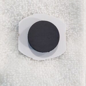 May include: A white plastic magnetic hook with a black round magnet. The hook is designed to hold items on a metal surface.
