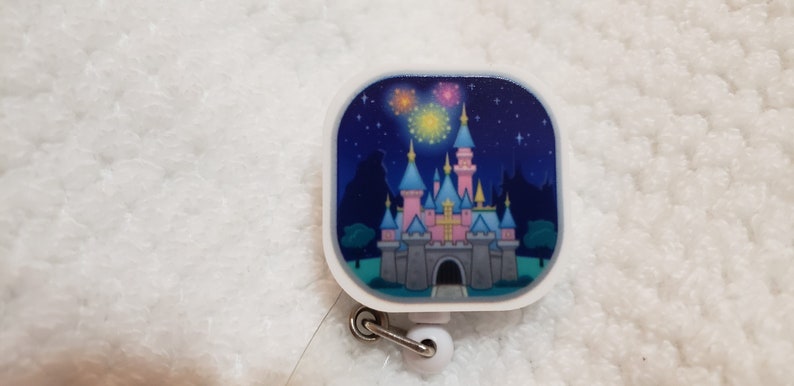 May include: A white retractable badge holder with a square-shaped charm featuring a cartoon image of Sleeping Beauty's castle with fireworks in the night sky.