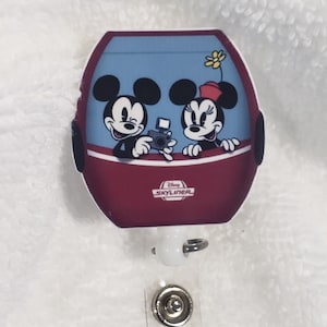 May include: A red and blue Disney Skyliner charm with Mickey and Minnie Mouse in the cabin. The charm is attached to a white retractable badge holder with two silver metal clips.