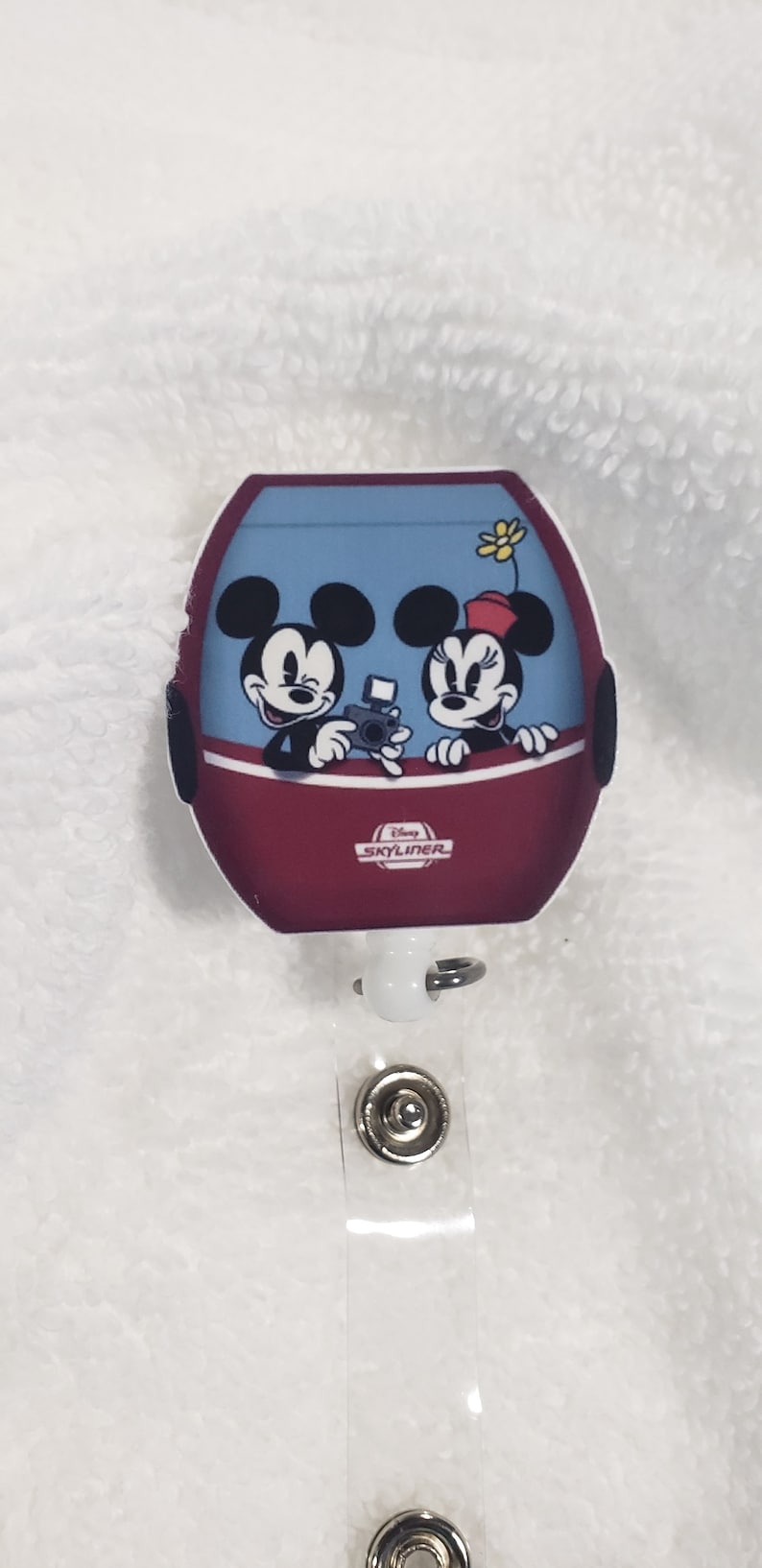 May include: A red and blue Disney Skyliner charm with Mickey and Minnie Mouse in the cabin. The charm is attached to a white retractable badge holder with two silver metal clips.