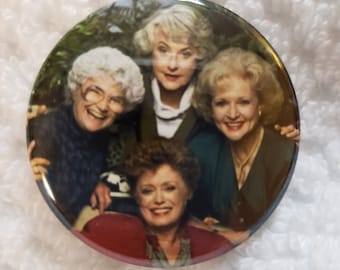 May include: A round button featuring a photo of four women, all smiling. The women are dressed in casual clothing and appear to be friends. The button is a light brown color with a glossy finish.