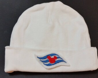 May include: A white beanie-style hat with a folded brim. The hat features a colorful emblem of a red Mickey Mouse head inside a blue and white stylized flag. The hat is made of a soft, plush material.
