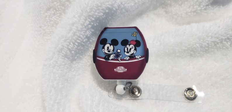 May include: A red and blue Mickey Mouse and Minnie Mouse themed retractable badge holder with a clear strap. The badge holder features Mickey and Minnie in a red and blue ride with the text "Disney's  Hollywood Studios" on the bottom.