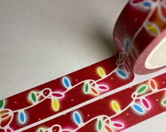 May include: Red washi tape with a repeating pattern of colorful Christmas lights. The tape is unrolled, showing the design clearly, with a roll of tape in the background. The lights are in shades of yellow, blue, green, and red.