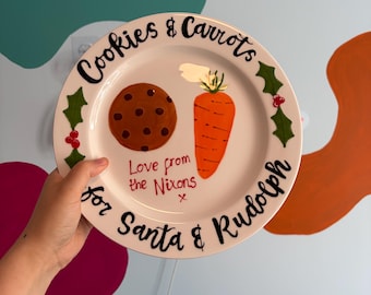 May include: A round white ceramic plate featuring a hand-painted brown chocolate chip cookie and an orange carrot. The text reads Cookies & Carrots for Santa & Rudolph, with Love from the Nixons written in red, accented by green holly leaves.