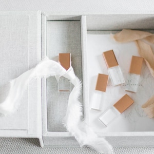 Può includere: Una scatola bianca con una fodera in tessuto e un nastro bianco. All'interno della scatola ci sono sei piccole unità USB rettangolari trasparenti con cime dorate.