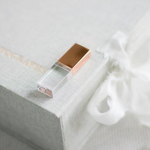 Może przedstawiać: Przezroczysty pendrive USB z miedzianą nasadką spoczywa na teksturowanej, jasnoszarej powierzchni. Pendrive znajduje się obok białej tkaniny o miękkiej, płynnej fakturze. Ogólna estetyka jest minimalistyczna i elegancka.