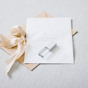 Op de afbeelding: Een witte envelop met een beige lint eromheen gebonden. Een kleine, transparante USB-stick met een zilveren bovenkant ligt op de envelop.