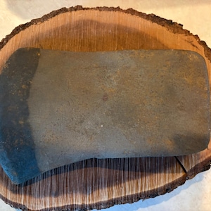 May include: A weathered, dark gray metal axe head with a wide, curved cutting edge and a rectangular poll. The surface of the metal shows signs of oxidation and age, with a textured, matte finish across the body and a darker, worn blade edge.