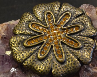May include: A large, ornate, bronze-colored metal belt buckle with a flower design. The flower has six petals and is decorated with small, amber-colored rhinestones.