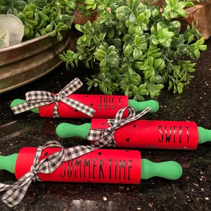 May include: Three red and green watermelon-themed rolling pins with black and white checkered ribbon bows. The rolling pins are decorated with black text that reads "Juicy", "Sweet", and "Summertime".
