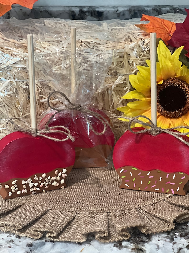 Pu&ograve; includere: Decorazioni in legno a forma di mela, con la parte superiore rossa e la parte inferiore color cioccolato, alcune con zuccherini. Le mele sono su una superficie di juta, con un girasole e foglie autunnali sullo sfondo. Ogni mela ha un bastoncino di legno.