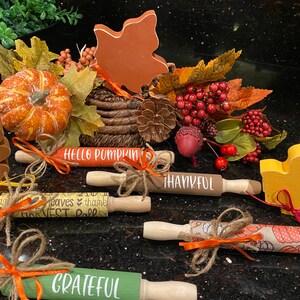 May include: A collection of fall-themed rolling pins with various designs and sayings, including "Hello Pumpkin", "Thankful", "Grateful", and "Leaves & Thanks Harvest Fall". The rolling pins are decorated with twine and orange ribbon. There are also wooden fall leaf decorations in orange, brown, and yellow.