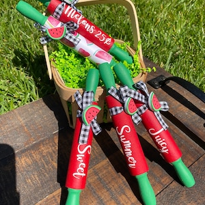 May include: Four red and green watermelon-themed rolling pins with white text. The rolling pins are in a basket with green shredded paper. The text on the rolling pins reads "Melons 25¢", "Sweet", "Summer", and "Juicy".