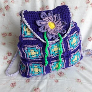 May include: Purple crocheted backpack with a flower design. The backpack has a green crocheted flower with a yellow center and purple petals. The backpack is made of squares with a light blue, yellow, and purple pattern.