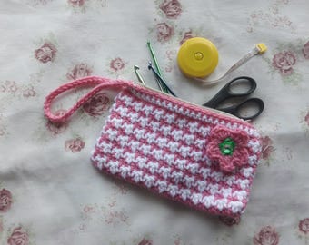 May include: A pink and white crocheted zippered pouch with a flower embellishment. The pouch is open and contains a tape measure, scissors, and crochet hooks.