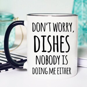 Puede incluir: Taza de cerámica blanca con asa y borde negros. La taza presenta el texto "DON'T WORRY, DISHES, NOBODY IS DOING ME EITHER" en negro y en negrita. Un artículo de cocina humorístico.