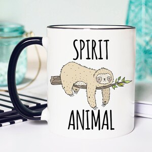 May include: White ceramic mug with a black handle. The mug has a black rim and features a cartoon sloth hanging from a branch with the text "SPIRIT ANIMAL" printed on the mug.