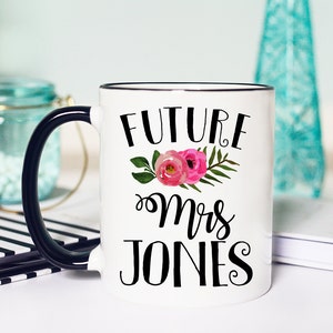 May include: White ceramic mug with a black rim and handle. The mug has black text that reads "FUTURE Mrs JONES" with a floral design in pink and green.