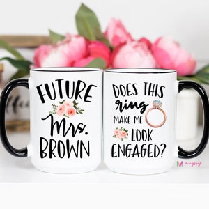 May include: Two white coffee mugs with black handles. The mug on the left says "Future Mrs. Brown" with a floral design. The mug on the right says "Does this ring make me look engaged?" with a diamond ring illustration.