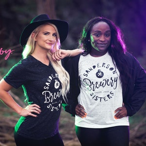 May include: Two women wearing t-shirts with the text "Sanderson Brewery Sisters" in a forest setting. One woman wears a black hat and a dark grey v-neck shirt. The other woman wears a white and grey t-shirt and a black jacket.