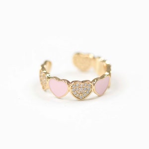 Puede incluir: Un anillo de oro con una serie de corazones de esmalte rosa, cada uno con una fila de piedras de circonita cúbica transparentes.