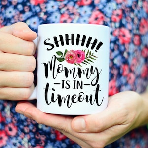 May include: White ceramic coffee mug with black text that reads "SHHHH! Mommy -is in- timeout" with pink flowers and green leaves.
