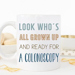 May include: White ceramic coffee mug with a blue and orange design. The mug reads "Look who's all grown up and ready for a colonoscopy."