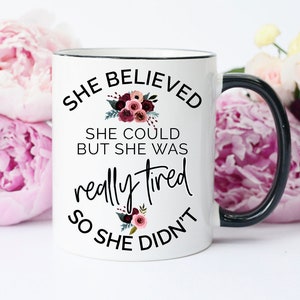 May include: White ceramic mug with a black handle and rim, featuring the text "She Believed She Could But She Was Really Tired So She Didn't." The design includes floral accents in shades of burgundy and green.