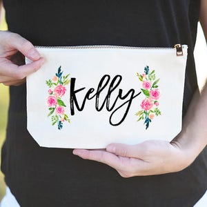 K&ouml;nnte beinhalten: Wei&szlig;e Kosmetiktasche aus Canvas mit Rei&szlig;verschluss. Die Tasche hat ein Blumenmuster mit rosa Bl&uuml;ten und gr&uuml;nen Bl&auml;ttern auf jeder Seite. Der Name "Kelly" ist in schwarzer Kursivschrift in der Mitte der Tasche geschrieben.