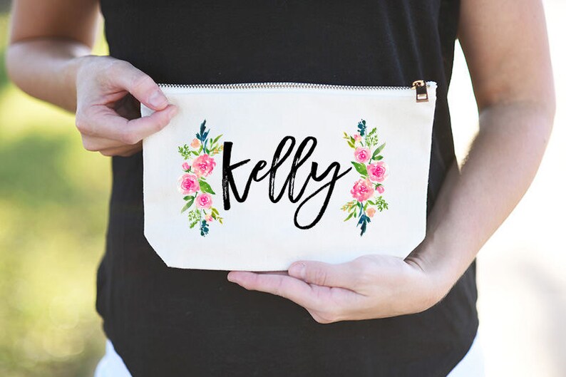 K&ouml;nnte beinhalten: Wei&szlig;e Kosmetiktasche aus Canvas mit Rei&szlig;verschluss. Die Tasche hat ein Blumenmuster mit rosa Bl&uuml;ten und gr&uuml;nen Bl&auml;ttern auf jeder Seite. Der Name "Kelly" ist in schwarzer Kursivschrift in der Mitte der Tasche geschrieben.