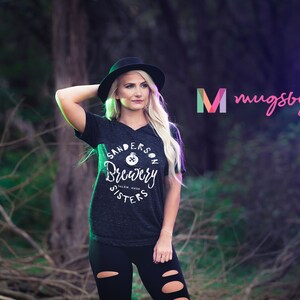 May include: Dark gray v-neck t-shirt with the text "Sanderson Brewery Salem Mass" in a circular design. The shirt is worn with black leggings and a black hat. The image also includes the logo "mugsby."