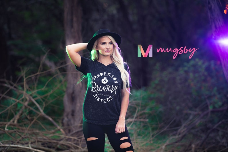 May include: Dark gray v-neck t-shirt with the text "Sanderson Brewery Salem Mass" in a circular design. The shirt is worn with black leggings and a black hat. The image also includes the logo "mugsby."