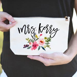 K&ouml;nnte beinhalten: Wei&szlig;e Kosmetiktasche aus Canvas mit Rei&szlig;verschluss. Die Tasche hat ein Blumenmuster und den Text "Mrs. Kerry" in schwarzer Schrift.