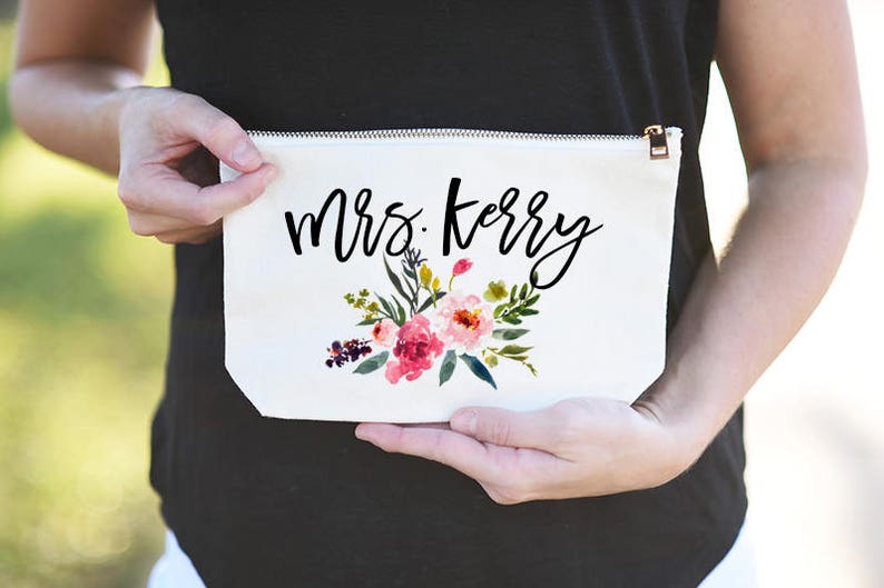 K&ouml;nnte beinhalten: Wei&szlig;e Kosmetiktasche aus Canvas mit Rei&szlig;verschluss. Die Tasche hat ein Blumenmuster und den Text "Mrs. Kerry" in schwarzer Schrift.
