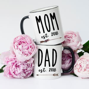 May include: Two white ceramic mugs with black handles and black lettering. The top mug says "MOM est. 2018" with a small arrow and floral design. The bottom mug says "DAD est. 2018" with a small arrow. The mugs are surrounded by pink peonies.