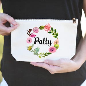 K&ouml;nnte beinhalten: Wei&szlig;e Kosmetiktasche aus Canvas mit einem floralen Kranzdesign und dem Namen "Patty" in schwarzen Buchstaben.