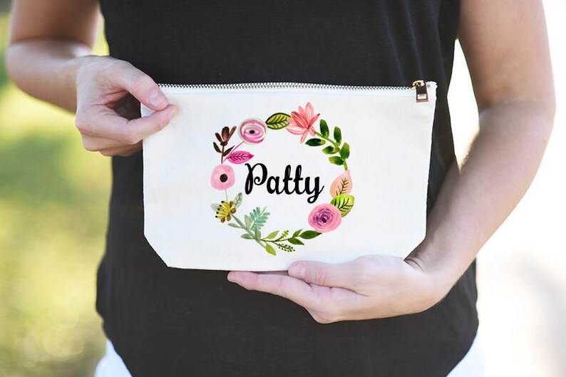 K&ouml;nnte beinhalten: Wei&szlig;e Kosmetiktasche aus Canvas mit einem floralen Kranzdesign und dem Namen "Patty" in schwarzen Buchstaben.