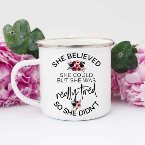 May include: White enamel mug with a silver rim and handle. The mug features the text "She Believed She Could But She Was Really Tired So She Didn't" in black script and a floral design. The mug is surrounded by pink flowers and green leaves.