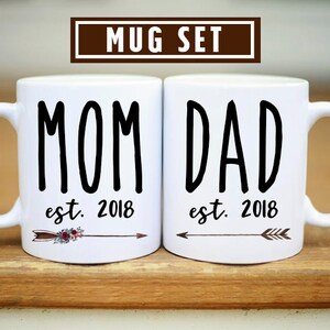 May include: White ceramic mugs with black text that reads "MOM est. 2018" and "DAD est. 2018". Each mug has a small arrow with floral accents.