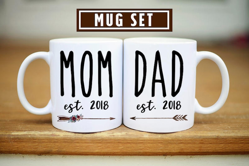 May include: White ceramic mugs with black text that reads "MOM est. 2018" and "DAD est. 2018". Each mug has a small arrow with floral accents.