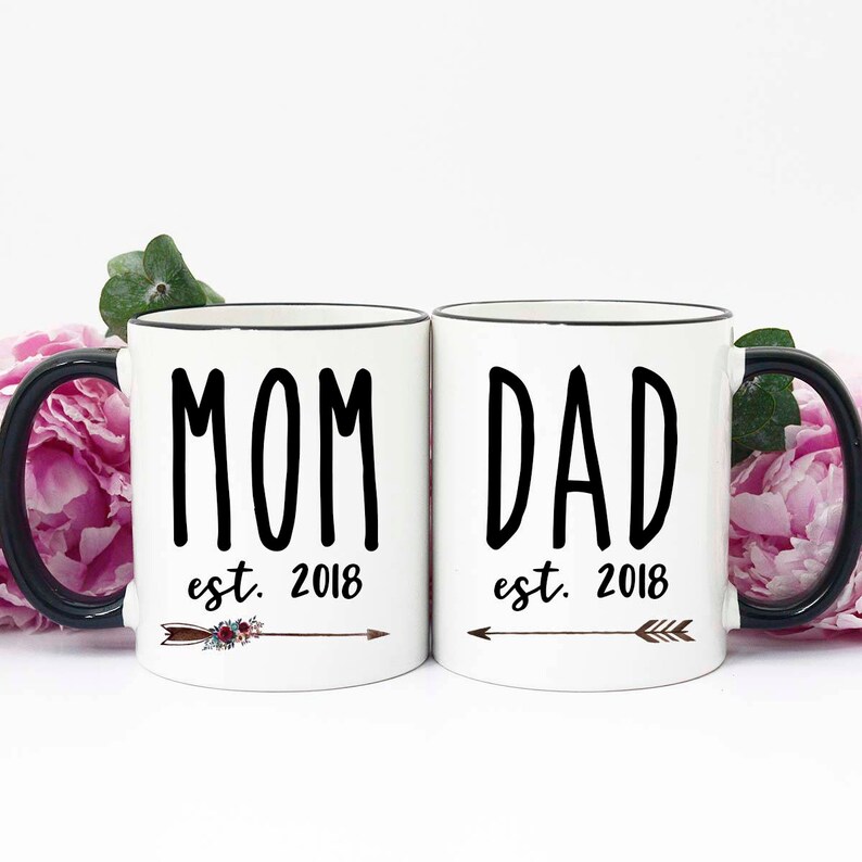 May include: Two white ceramic mugs with black rims and handles. The mug on the left says "MOM est. 2018" with a brown arrow and floral design. The mug on the right says "DAD est. 2018" with a brown arrow.