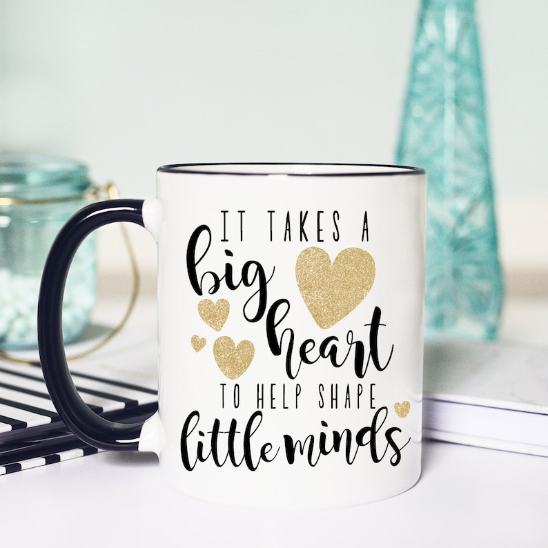 May include: White ceramic coffee mug with a black rim and handle. The mug has a black and gold glitter design with the text "It takes a big heart to help shape little minds".
