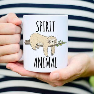 May include: White ceramic mug with a black outline of a sloth hanging from a branch. The mug has the text "SPIRIT ANIMAL" printed on it.