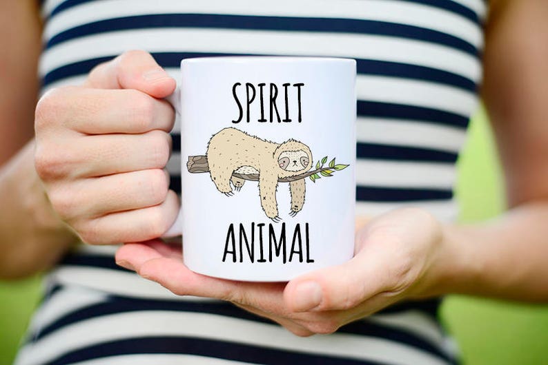 May include: White ceramic mug with a black outline of a sloth hanging from a branch. The mug has the text "SPIRIT ANIMAL" printed on it.