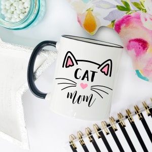 May include: White ceramic mug with a black handle and black rim. The mug has a black and white illustration of a cat face with pink ears and a pink heart nose. The text "CAT MOM" is written below the cat face.